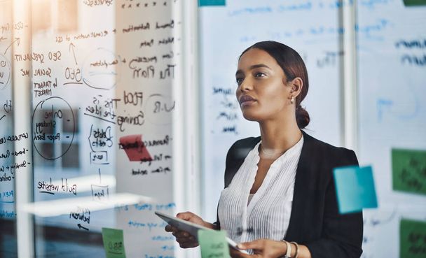 She sets out her goals very meticulously. a young businesswoman using a digital tablet while brainstorming notes on a glass wall in an office - Photo, Image