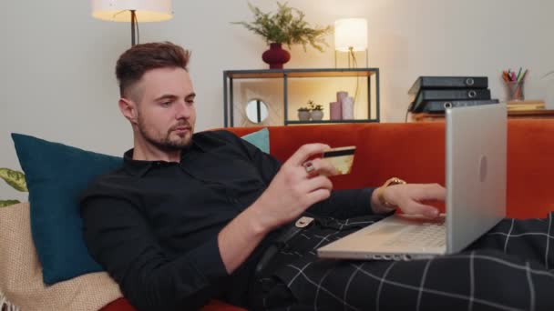 Man using credit bank card and laptop computer while transferring money, purchases online shopping, order food delivery at home. Young guy sitting on couch holding dollar cash at home living room - Footage, Video