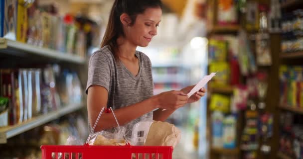 Nuori vaimo tai äiti käyttää ostoslistaa ostaakseen ostoksia kaupasta. Nainen kaupan käytävällä ruokakorin kanssa etsimässä esinettä. 4 k. - Materiaali, video
