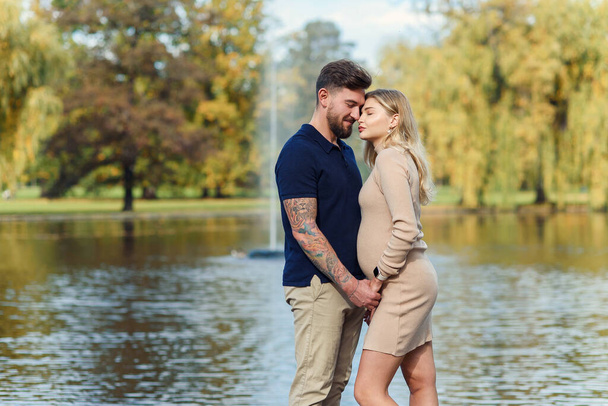 Stylish young pregnant woman and her handsome husband hug each other at the autumn park against the lake. - Φωτογραφία, εικόνα