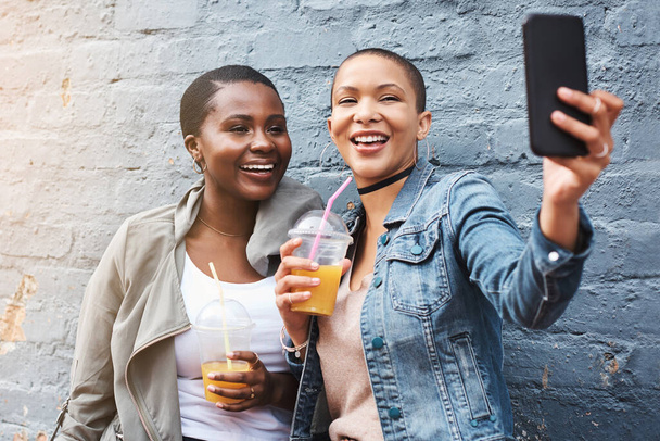 Teilen Sie jeden Moment, während Sie mitgehen. zwei junge Frauen stehen lächelnd neben einem Gebäude und machen Selfies, während sie ihre kühlen Drinks halten - Foto, Bild