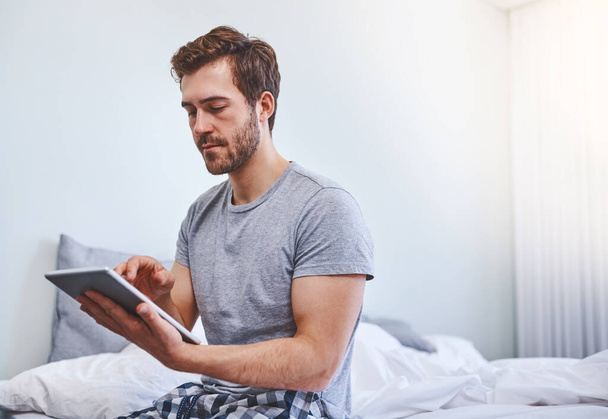 O dispositivo ajuda-o a planear o dia. um jovem bonito usando um tablet enquanto sentado em uma cama em casa - Foto, Imagem