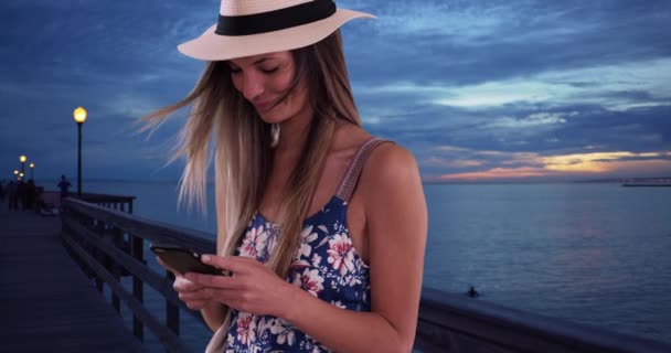 Gelukkige vrouw in bloemen romper en fedora sms 'en op de pier in de avond. Millennial meisje in haar twintiger jaren sms 't op een mobieltje buiten bij de oceaan. 4k - Video