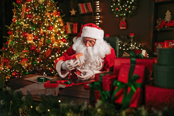 Santa Clause is packing gifts while sitting at the table near Christmas tree at home. New Year concept - Photo, image