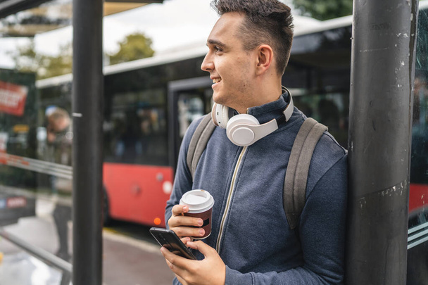 Um homem jovem adulto masculino stand na estação de ônibus de transporte público esperando com fones de ouvido e telefone inteligente móvel no inverno ou no outono dia com mochila estudante ou turista cidade vida cópia espaço - Foto, Imagem