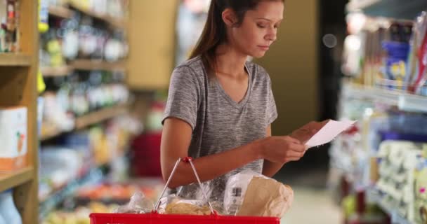 Mooie jonge vrouw in de supermarkt die haar boodschappenlijstje bekijkt. Jonge vrouw of moeder klant dragen mand op zoek naar item in de winkel gangpad. 4k - Video