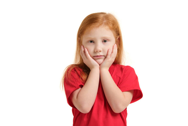 Szomorú szürke szemű kislány portréja, kezével az arcán, fehér háttérrel elszigetelve. Egy vörös pólós lány érzelmei. - Fotó, kép