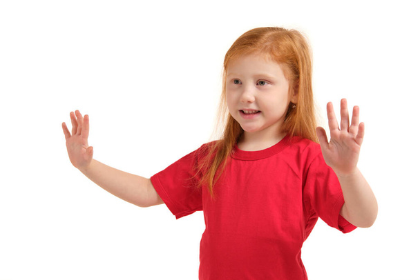 A kislány feltartott karokkal mosolyog. 5 éves lány meglepett mosollyal és a kezét a kezét. Vörös hajú lány piros pólóban elszigetelt fehér háttér - Fotó, kép