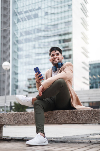 νεαρός Αμερικανός άνδρας που χρησιμοποιεί smartphone κάθεται σε εξωτερικούς χώρους - Φωτογραφία, εικόνα