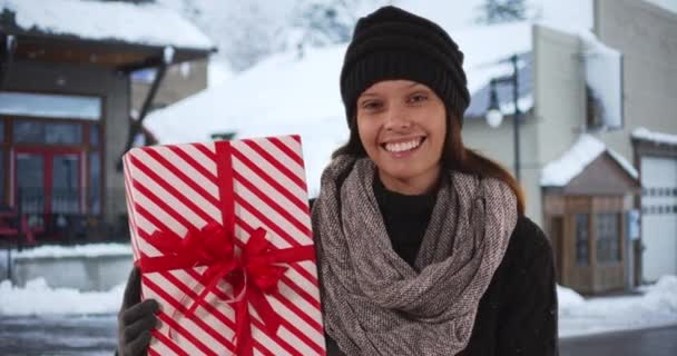 Закрыть портрет женщины, улыбающейся и держащей рождественский подарок на снежной улице в центре города. Счастливая миллениалка с праздником смотрит в камеру. 4k - Кадры, видео