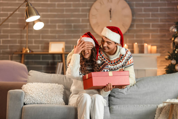 Молодой человек удивляет свою любимую девушку подарком дома в канун Рождества - Фото, изображение