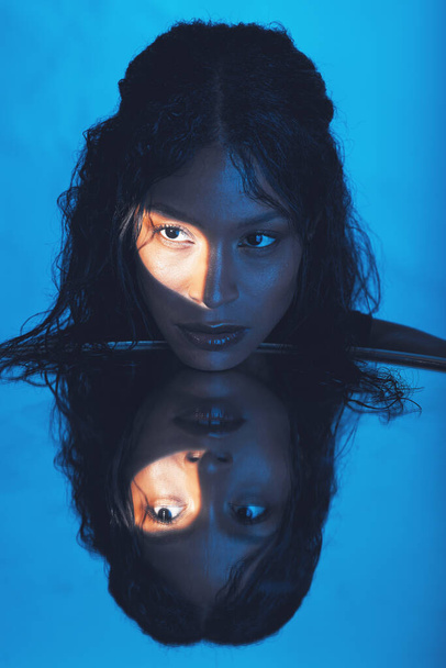 Beauty, woman and light on face reflection with mirror for edgy cosmetics, skincare or advertising. Shadow, thinking and spotlight on beautiful cosmetic black model with blue studio background - 写真・画像