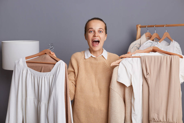 Indoor shot of excited amazed young adult woman holding clothing on two hangers in boutique, choosing between two looks, expressing excitement, yelling happily. - Foto, imagen