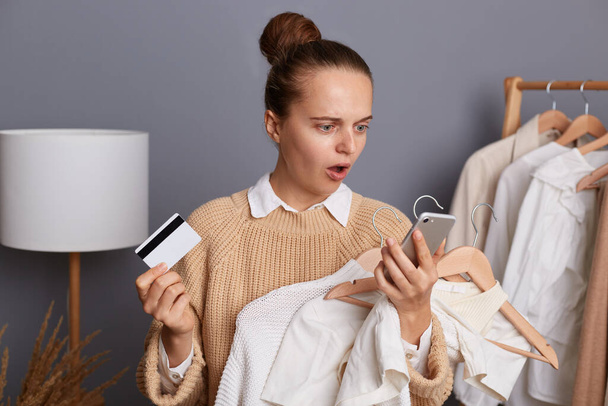 Portrait of shocked astonished young adult woman holding clothing on hangers in shopping mall, holding mobile phone and credit card in hands, looking at screen of phone, sees she has no money. - Foto, afbeelding
