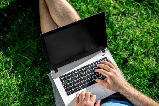 Cropped shot of man using laptop with blank screen while sitting on green grass. Freelance works in the open air in the park - Foto, Bild