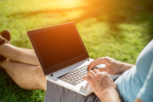 Cropped shot of man using laptop with blank screen while sitting on green grass. Freelance works in the open air in the park. Sun flare - Fotografie, Obrázek