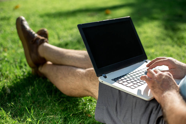 Καλλιεργημένη φωτογραφία του ανθρώπου που χρησιμοποιεί φορητό υπολογιστή με κενή οθόνη, ενώ κάθεται στο πράσινο γρασίδι. Ελεύθερος επαγγελματίας εργάζεται στην ύπαιθρο στο πάρκο - Φωτογραφία, εικόνα