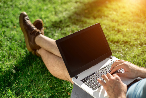 Cropped shot of man using laptop with blank screen while sitting on green grass. Freelance works in the open air in the park. Sun flare - Фото, зображення