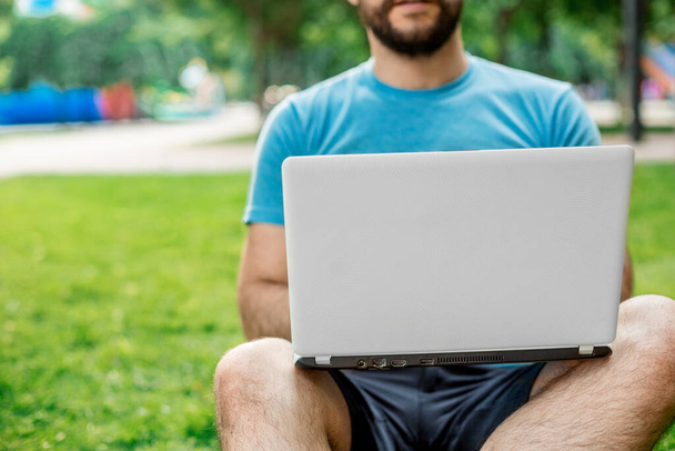 Молодой человек использует и печатает ноутбук в летней траве. Фрилансер, работающий в открытом парке - Фото, изображение