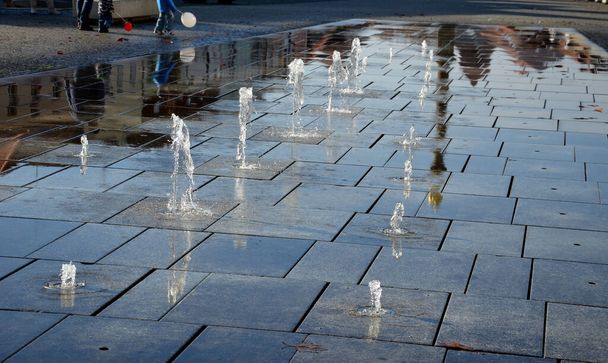 Quadratische Granitpflastersteine stapeln sich in einem Streifen auf dem Bürgersteig, Junge, Wasserspritzer mit wechselnden Sprühhöhen. Wasserspiele entstehen direkt aus dem gepflasterten Platz in der Parkreihe. nasse Fliesen Promenade, Baby, Mütze - Foto, Bild