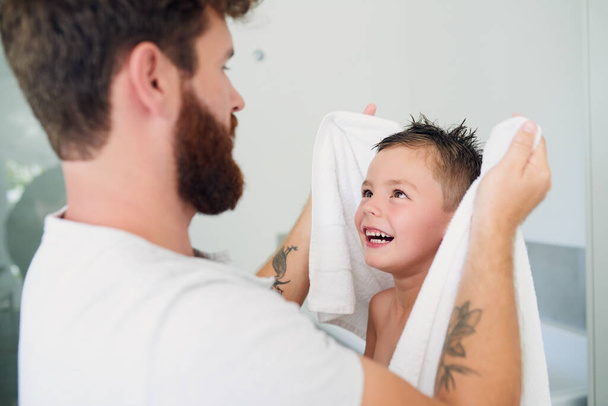 Όμορφα και καθαρά, τώρα για να στεγνώσουμε. Ένας νεαρός όμορφος πατέρας στεγνώνει τον αξιολάτρευτο γιο του μετά από ένα μπάνιο στο σπίτι. - Φωτογραφία, εικόνα