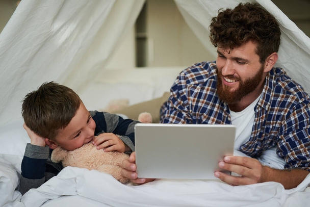 Şuna bir bak. Yakışıklı bir genç adam ve oğlu evdeki yatakta yatarken tablet kullanıyorlar. - Fotoğraf, Görsel