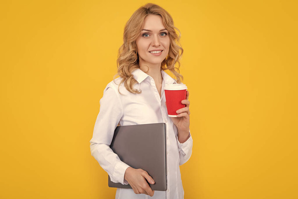 Sarı arka planda kahve fincanı ve dizüstü bilgisayarı olan gülümseyen sarışın kadın.. - Fotoğraf, Görsel