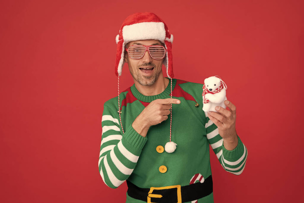 Erstaunter Mann mit Parteibrille zeigt mit dem Finger auf Weihnachtsdekoration Schneemann auf rotem Hintergrund. Frohes neues Jahr. Frohe Weihnachten. Mann im Elfenkostüm und Weihnachtsmann-Hut. - Foto, Bild