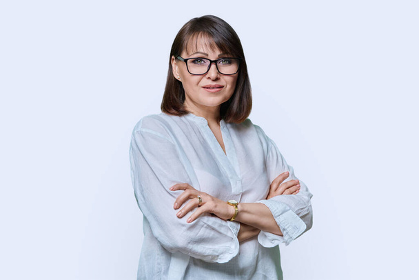 Positive smiling confident middle aged woman looking at camera on white studio background. Friendly beautiful mature female with hairstyle makeup wearing glasses, white shirt with crossed arms. - Fotografie, Obrázek