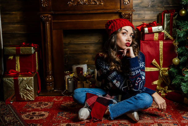 Žena v červené čepici a šátku se uraženě zamyslela. Krásná bruneta sedí pod vánočním stromečkem na pozadí novoroční výzdoby. - Fotografie, Obrázek