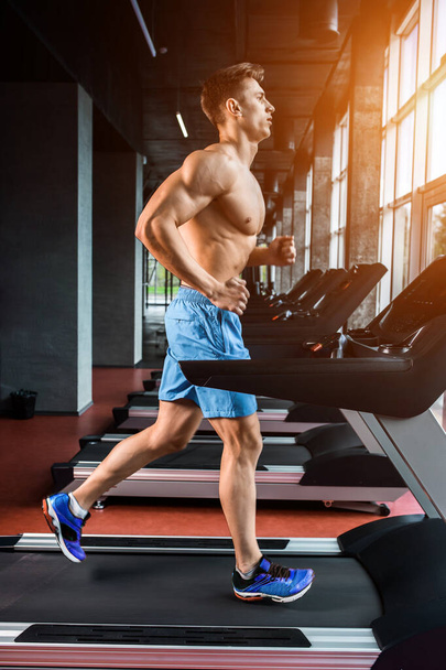 Side view full length of young man in sportswear running on treadmill at gym. Muscular young man in blue shorts doing exercises. sun flare - Photo, Image