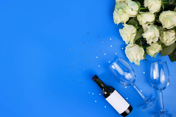 Top view photo of romantic evening, dinner decor hearts love wineglasses with confetti wine bottle and white tape with large bouquet of white roses on isolated blue background with copyspace - Foto, afbeelding