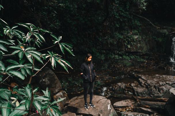 barna kaukázusi lány fekete kabát sötét nadrág és tornacipő fókuszon kívül nyugodt és nyugodt megcsodálva a szépség vízesések és erdei növényzet, purakaunui esik, új-Zéland - utazási koncepció - Fotó, kép