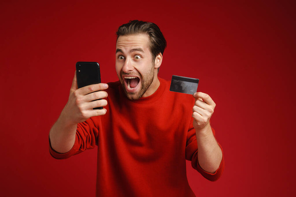 Young shocked man screaming while using cellphone and credit card isolated over red background - Zdjęcie, obraz