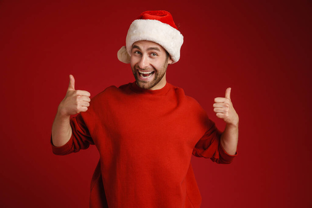 Young white man wearing santa hat smiling and showing thumbs up isolated over red background - Фото, изображение