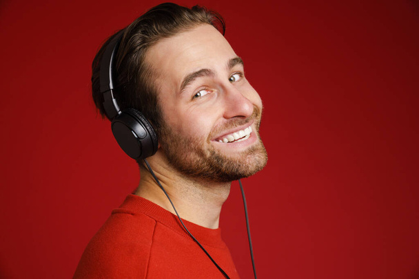 Young white man smiling while listening music with headphones isolated over red background - 写真・画像