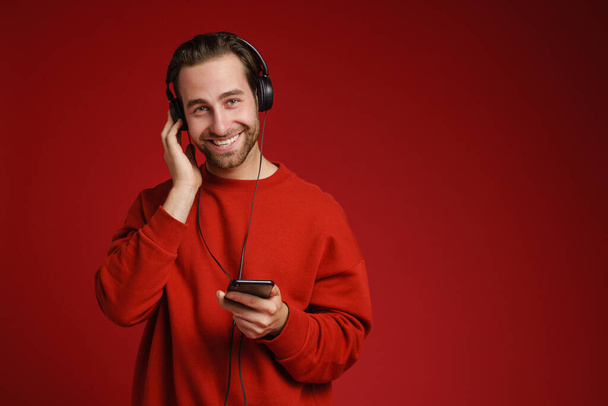 Junger weißer Mann lächelt beim Musikhören mit Handy isoliert über rotem Hintergrund - Foto, Bild