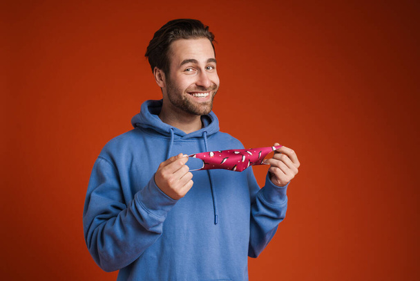 Young bristle man smiling while posing with face mask isolated over red background - 写真・画像