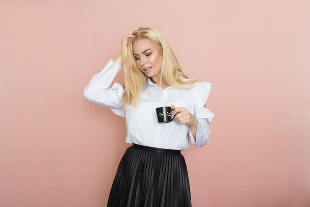 Beauty, fashion portrait. Elegant business style. Portrait of a beautiful blonde woman in white blouse and black skirt posing at studio on a pink background. Holding black cup in her hands. Drinking coffee, tea - Zdjęcie, obraz