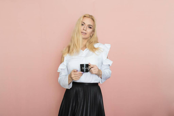 Beauty, fashion portrait. Elegant business style. Portrait of a beautiful blonde woman in white blouse and black skirt posing at studio on a pink background. Holding black cup in her hands. Drinking coffee, tea - Фото, изображение