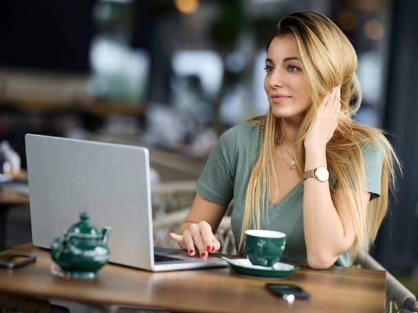 Ein lächelnder Freiberufler sitzt in einem Café und tippt auf einem Laptop, während er auf den Monitor schaut. - Foto, Bild