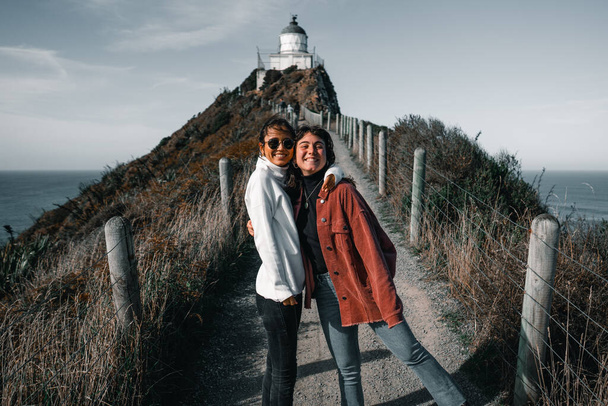 Bir çift beyaz kadın kameraya bakıyor mutlu ve mutlu bir şekilde kucaklaşıyorlar dar çitli yolda deniz fenerine, Nugget Point 'e, Yeni Zelanda' ya doğru yol alıyorlar. - Fotoğraf, Görsel