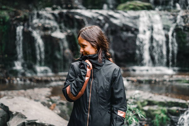 ragazza caucasica con gli occhi neri e capelli castani felice e calma tenendo la cerniera della sua giacca nera dalle cascate e lago in mezzo alla natura, purakaunui cade, Nuova Zelanda - concetto di viaggio - Foto, immagini