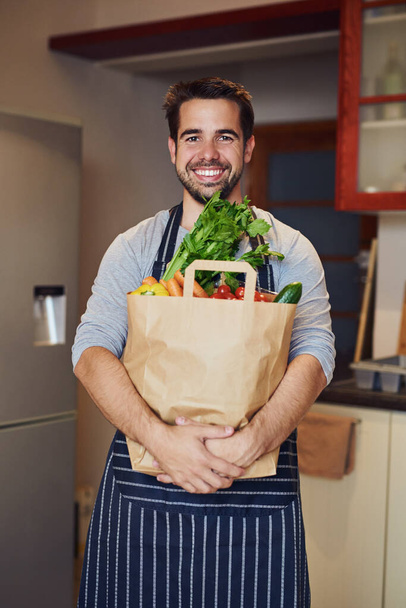 Všechna má jídla začínají výjimečnými ingrediencemi. Portrét šťastného mladého muže pózujícího s taškou potravin v kuchyni doma - Fotografie, Obrázek