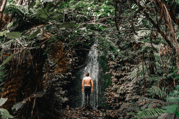 Niño caucásico con el pelo largo atado hacia atrás, espalda desnuda y pantalones negros de pie sobre una roca viendo caer el agua de la cascada en el exuberante bosque en el tranvía waitawheta, Nueva Zelanda - Estilo de vida - Foto, imagen