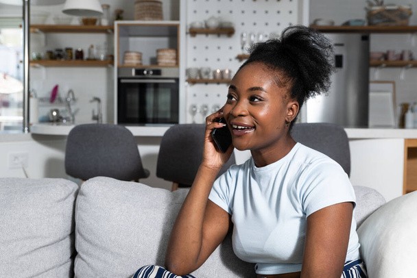 Pozitif genç, gururlu siyahi bayan cep telefonuyla sohbet ediyor ve evdeki kanepede gülümsüyor. Neşeli Afro-Amerikan kadın kapalı alanda akıllı telefondan biriyle iletişim kuruyor. - Fotoğraf, Görsel
