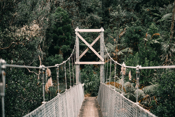 długi wąski stary most z filarami i drewnianą podłogą zapinane długimi metalowymi drutami, które przekraczają rzekę lasu na wyspie pączków, Nowa Zelandia - koncepcja stylu życia - Zdjęcie, obraz