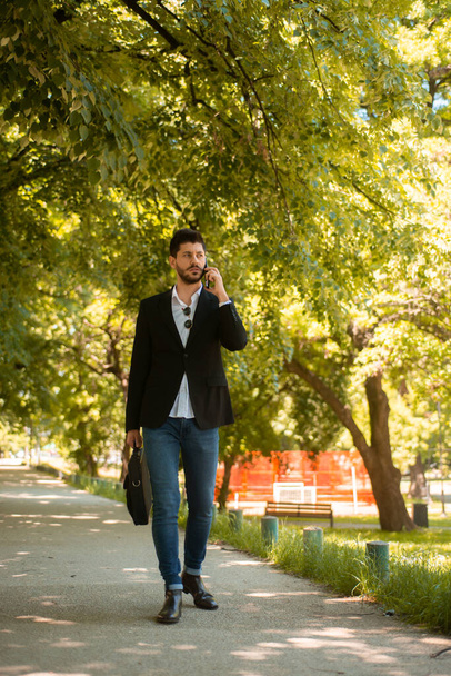 Young handsome bussinessman is discussing something important on the phone while walking in the park - 写真・画像