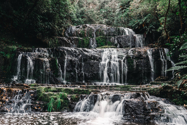 cascade d'eau se précipitant dans le lac parmi la mousse et l'herbe par les rochers impressionnants au milieu de la verdure près des arbres forestiers, chutes purakaunui, Nouvelle-Zélande - Concept Voyage - Photo, image