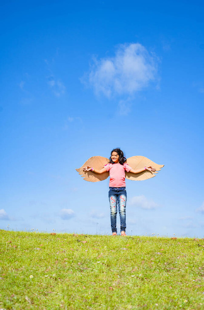Vertikale Aufnahme, fröhliches aufgeregtes Mädchen mit künstlichen Flügeln, das wie ein Vogel auf einem Hügel in grüner Wiese vor blauem Himmel tanzt - Konzept von Freiheit, Glück und Unabhängigkeit. - Foto, Bild